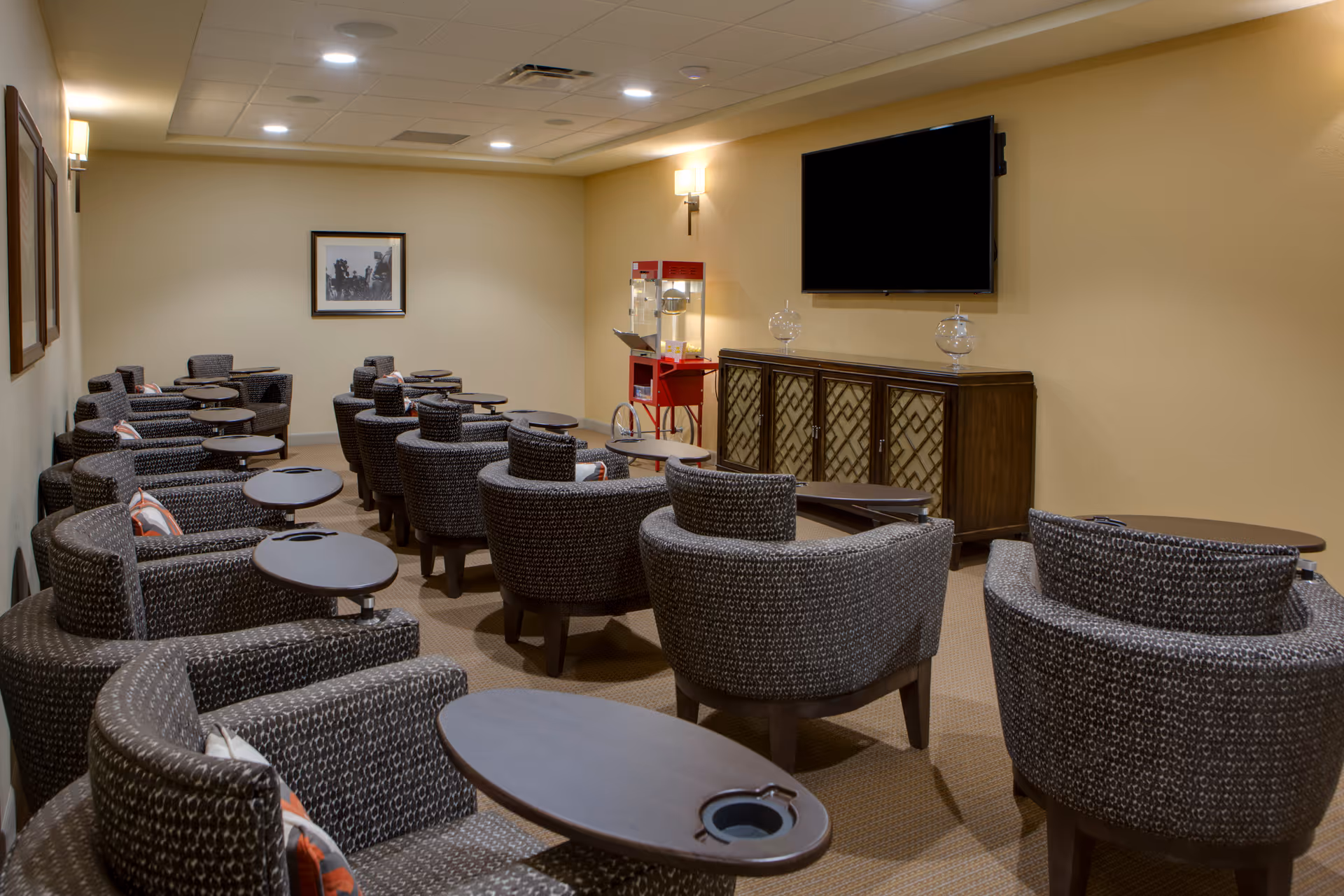A small theater-style room with multiple rows of cushioned armchairs, each equipped with a small attached table. At the front of the room is a large flat-screen TV mounted on the wall above a wooden cabinet. A red popcorn machine is positioned in the corner near the TV. The walls are painted beige and decorated with framed pictures, and the room is softly lit with wall sconces.