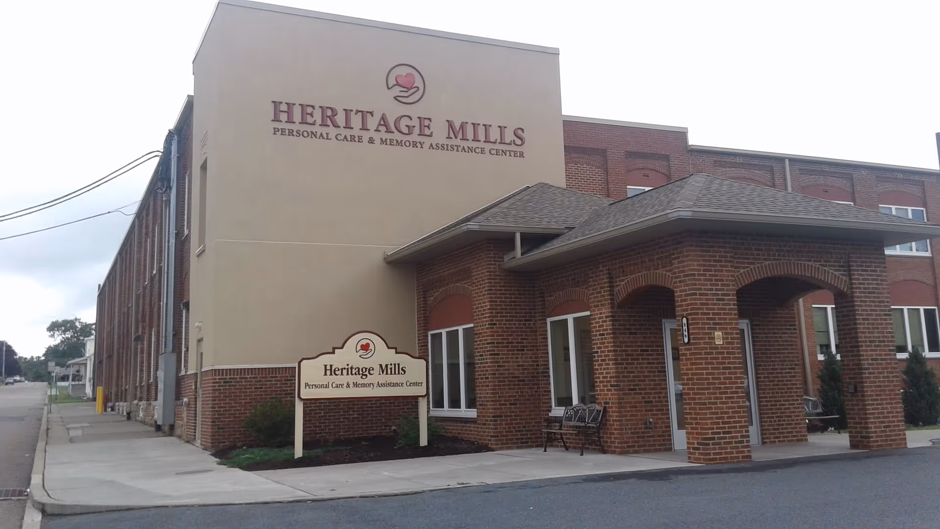 Exterior view of Heritage Mills Personal Care & Memory Assistance Center, showing a brick building with a covered entrance and signage on the building and a freestanding sign near the sidewalk.
