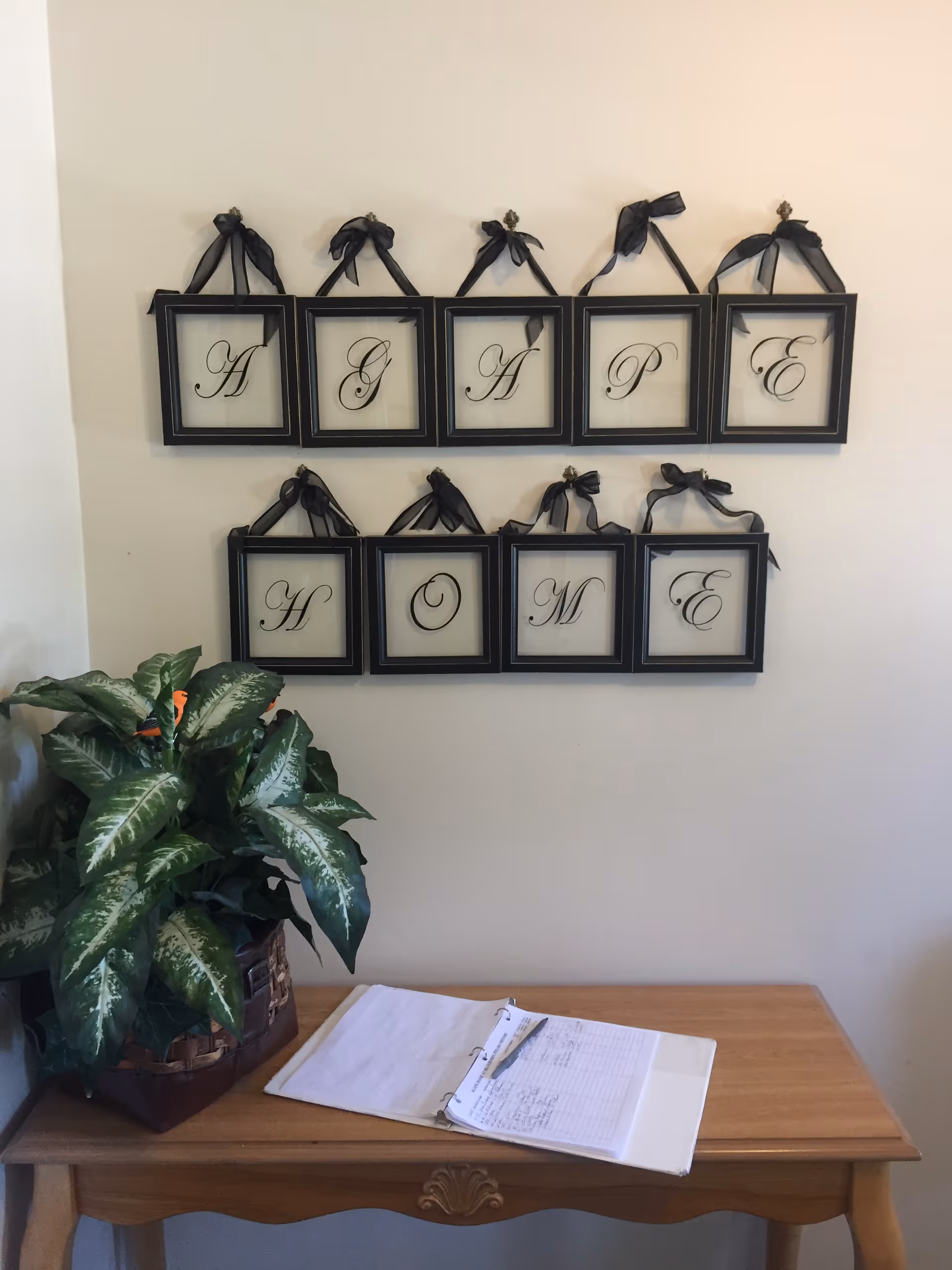 A decorative wall display with nine black-framed letters spelling 'AGAPE HOME' arranged in two rows, hanging by black ribbons on a beige wall above a wooden table. On the table, there is a green leafy potted plant and an open binder with a pen resting on it.