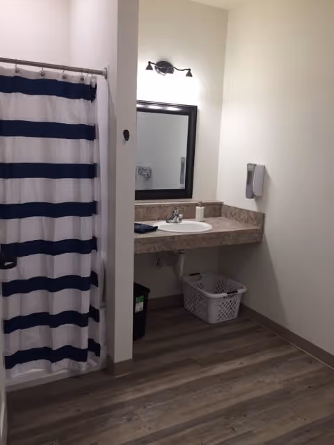 A clean bathroom with a navy-and-white striped shower curtain, sink and mirror on a stone countertop, wall-mounted soap dispenser and a laundry basket on wood-look flooring.