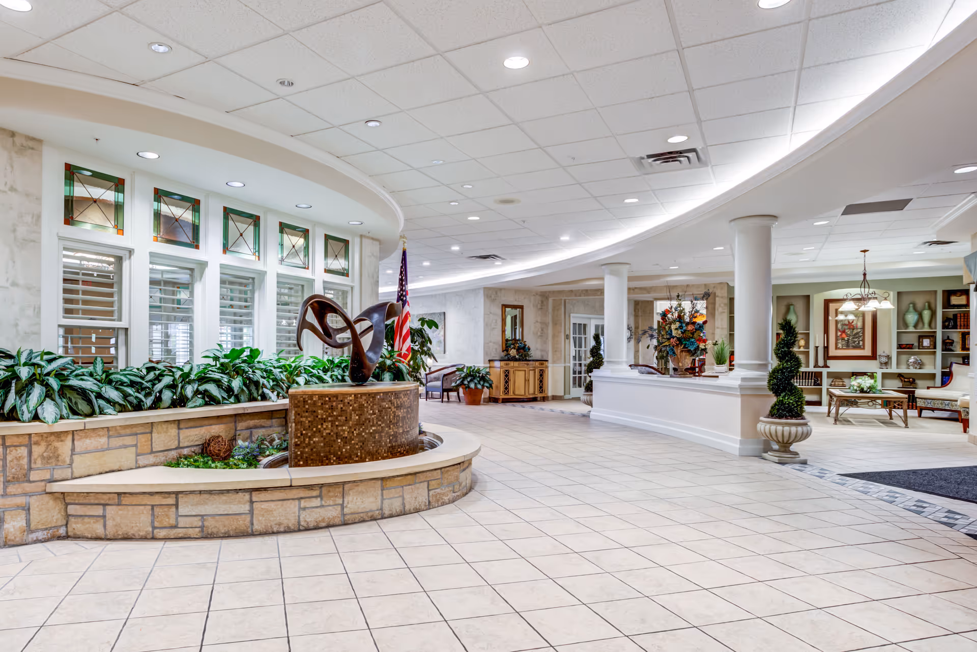 Spacious and well-lit interior lobby area with tiled floors, a curved stone planter filled with green plants, and a modern abstract sculpture in the center. The space features white columns, decorative stained glass windows, an American flag, and comfortable seating areas with floral arrangements and framed artwork.