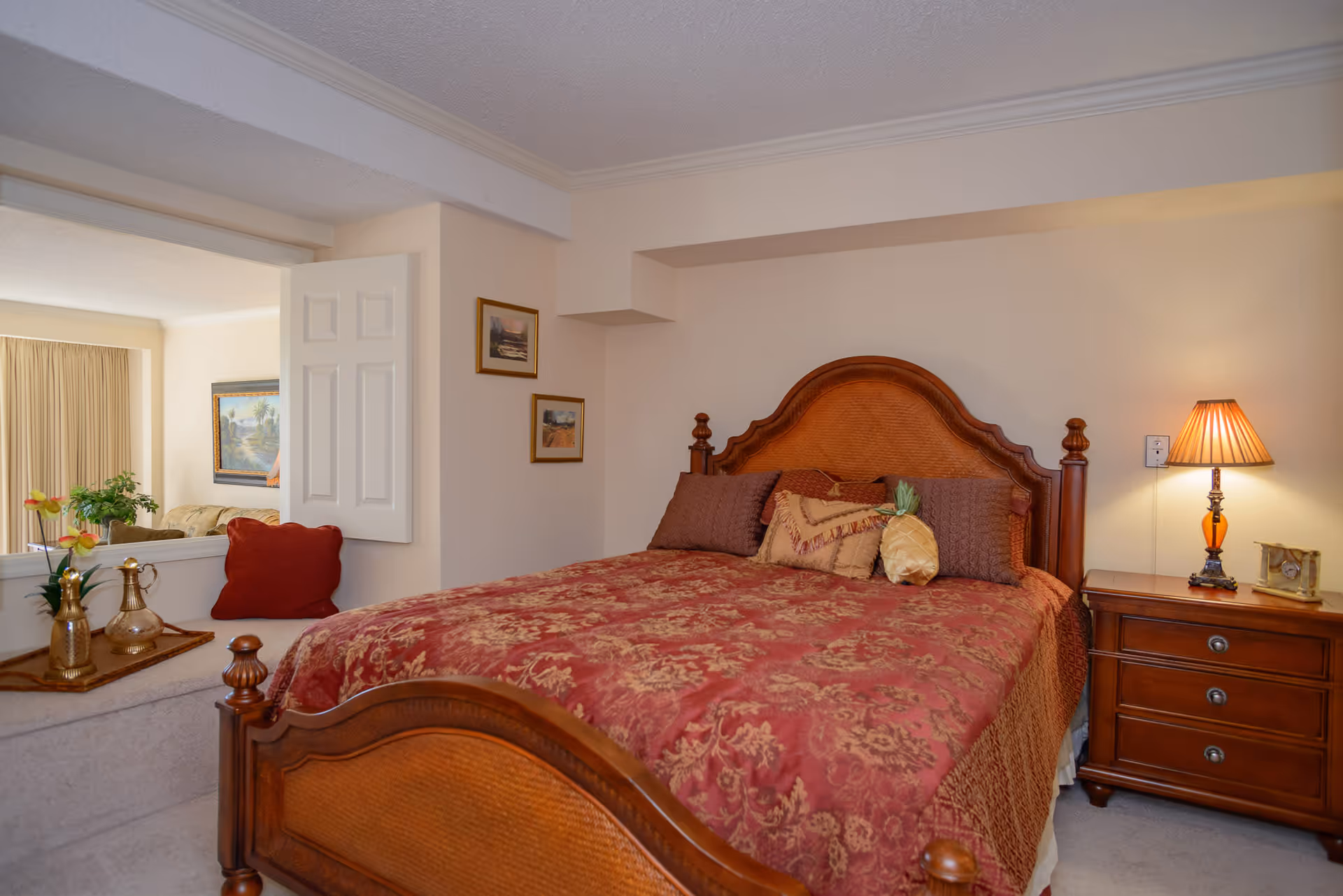 A bedroom with a wooden bed frame and a red and gold patterned bedspread. The bed has multiple pillows, including one shaped like a pineapple. There is a wooden nightstand with a lamp and a small clock on it. On the left side, there is a window-like opening showing a glimpse of a living room with a sofa, a plant, and framed artwork on the walls.