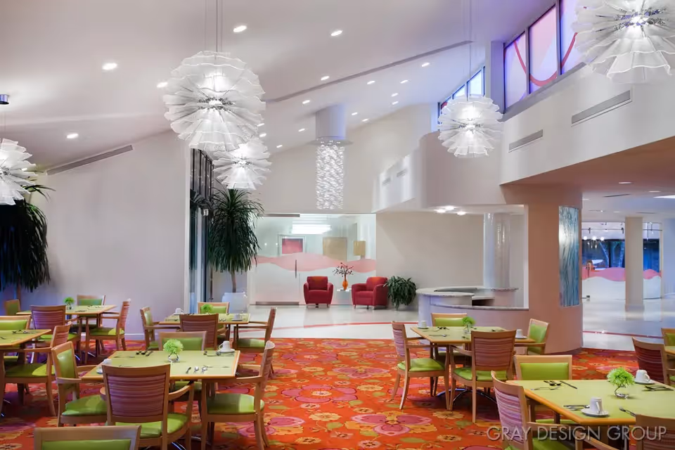 Spacious, modern dining room with green-topped tables and chairs, a red floral carpet, large pendant lights, and a seating area in the background.