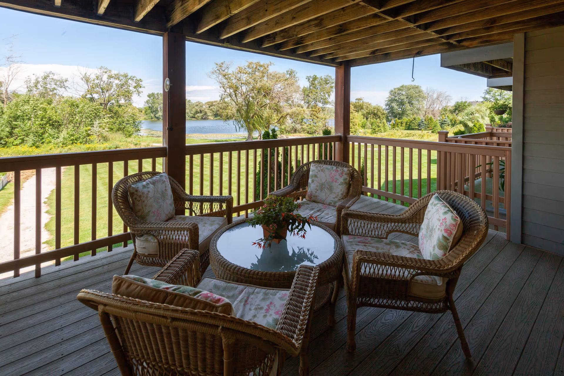 Covered wooden porch with wicker seating and a glass-topped table overlooking a grassy lawn and lake.
