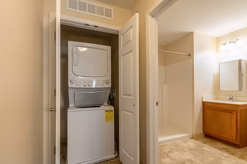 Stacked washer and dryer in an open closet next to a bathroom with a shower, vanity, and mirror.