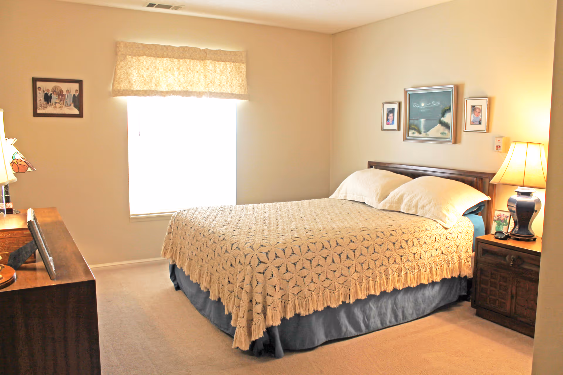 A cozy bedroom with a large bed covered by a cream-colored crocheted blanket and two pillows. There is a wooden nightstand with a lamp and a small picture frame on the right side of the bed. On the left side, there is a wooden dresser with a lamp and some decorative items. The room has beige walls with three framed pictures hanging above the bed and one framed picture on the wall next to the window, which is covered by a beige valance.