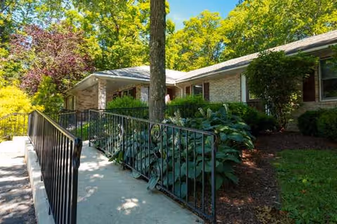 Exterior view of a single-story brick building surrounded by lush green trees and shrubs, with a concrete walkway and black metal railing leading to the entrance.