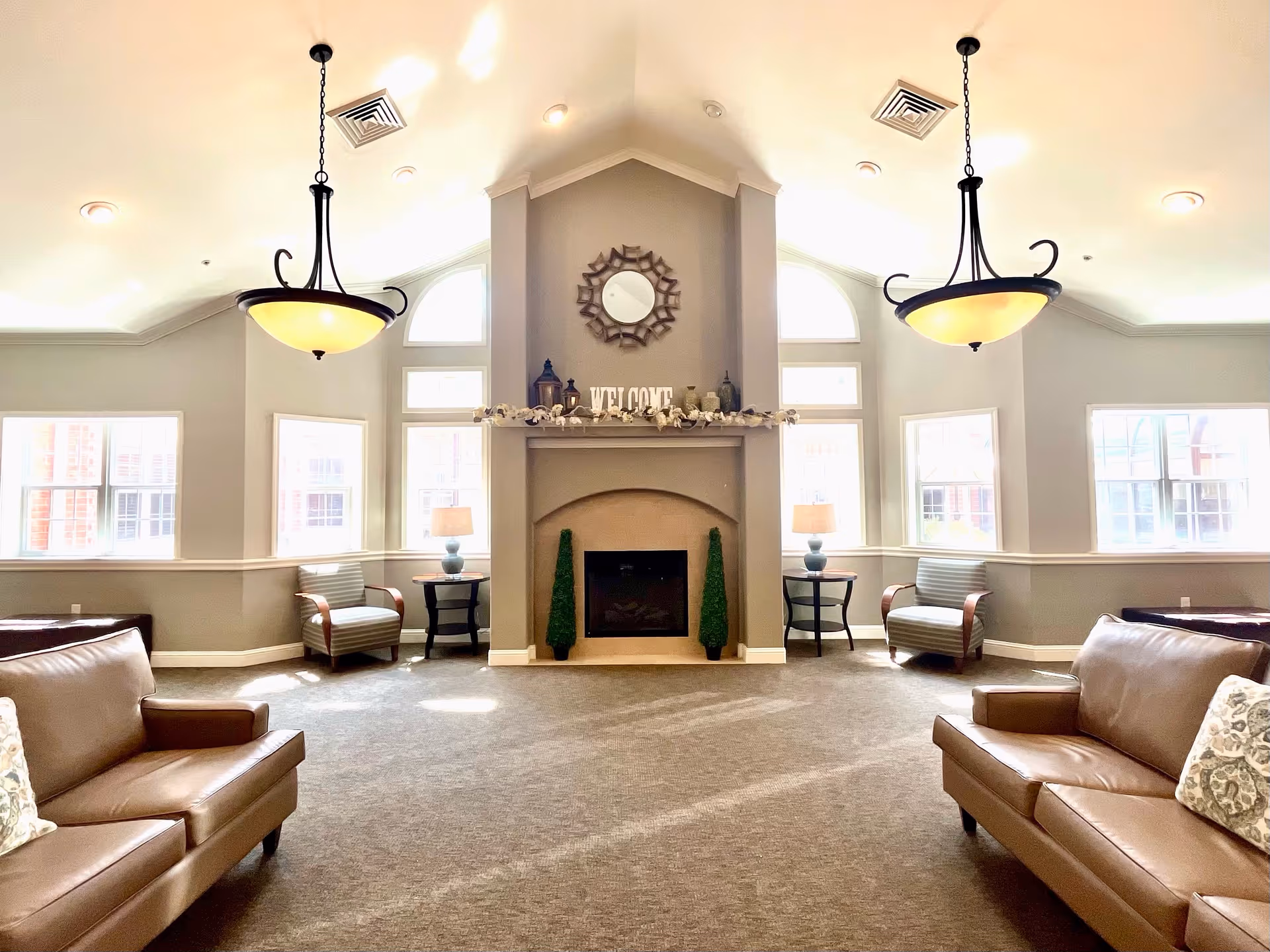 A bright and spacious living room with high vaulted ceilings, two hanging light fixtures, a central fireplace decorated with a mirror, lanterns, and a 'WELCOME' sign. The room features large windows letting in natural light, two armchairs with side tables and lamps, and two brown leather sofas with patterned pillows.