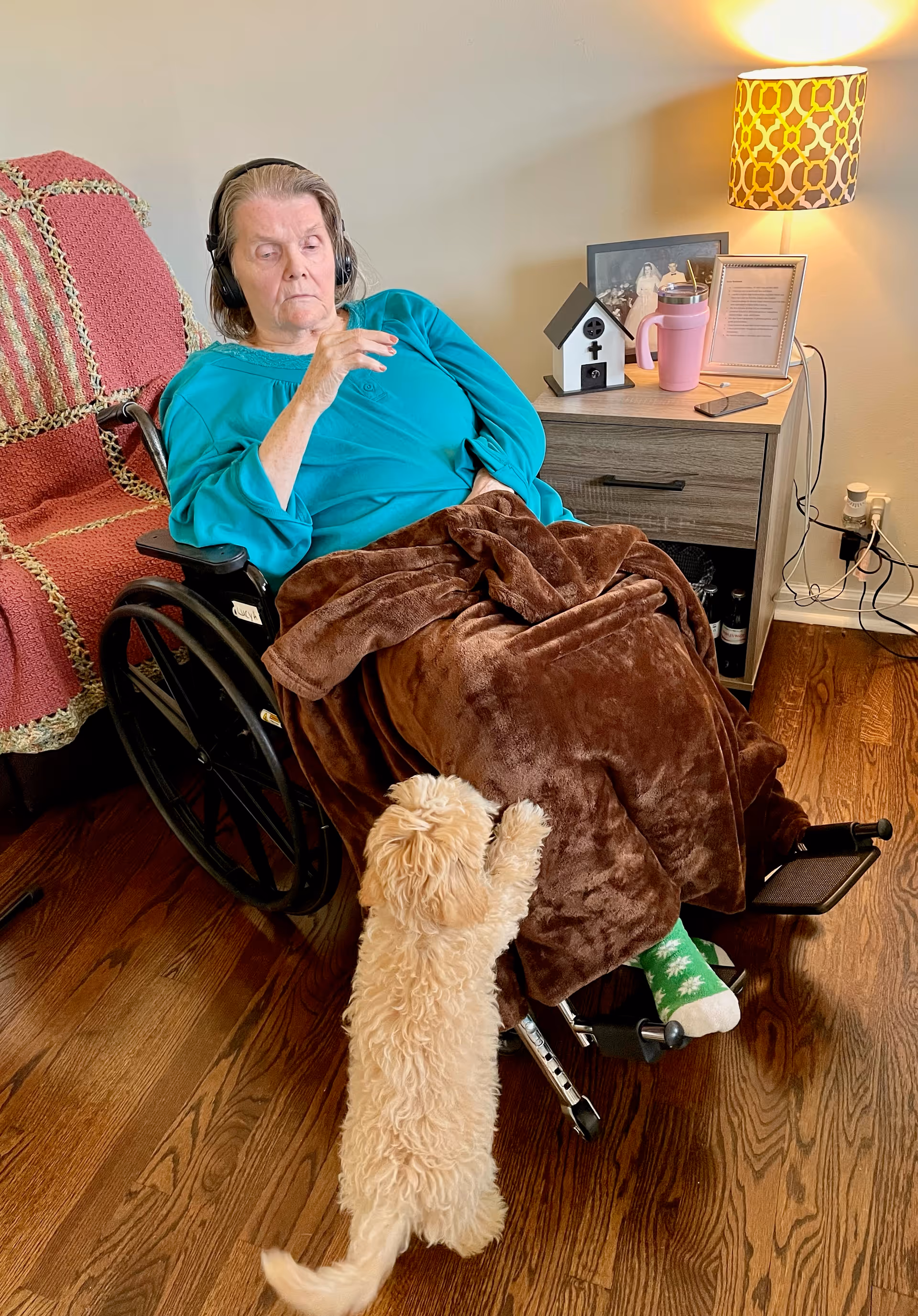 A woman wearing headphones sits in a wheelchair covered by a brown blanket while a small dog stands on its hind legs at her lap next to a side table with a lamp.