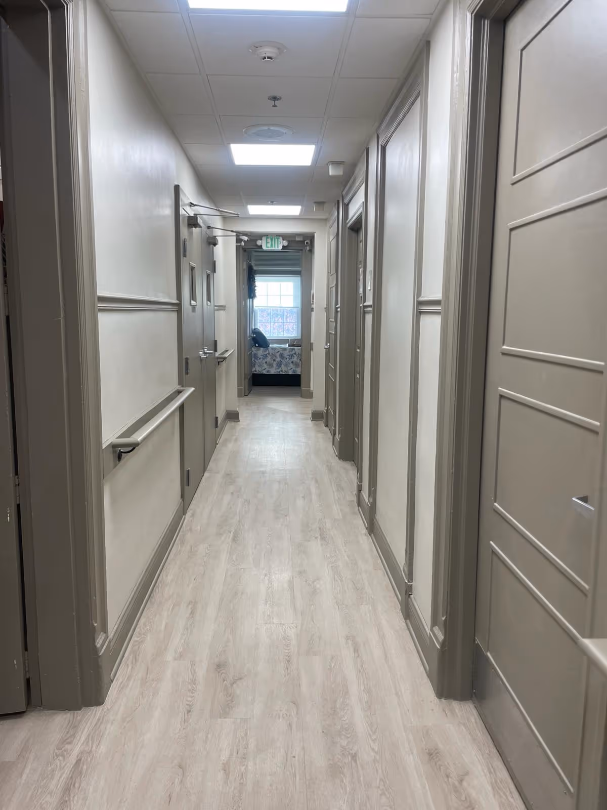 A long, well-lit hallway in an assisted living facility with light wood flooring and beige walls. Several closed doors line both sides of the hallway, and a handrail runs along the left wall. At the end of the hallway, a window with natural light and a bed with pillows are visible.