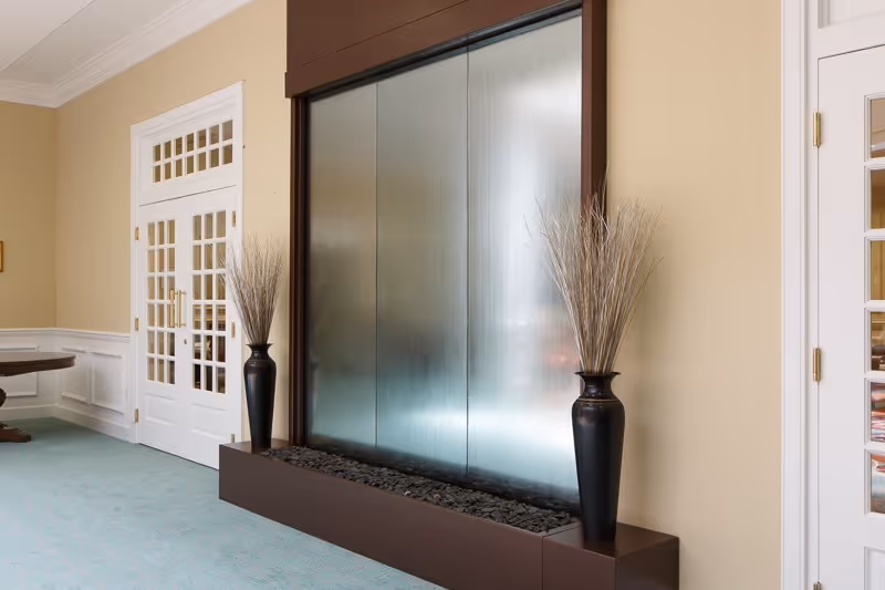 Interior common area with a tall glass water wall flanked by decorative vases and glass-paneled doors.