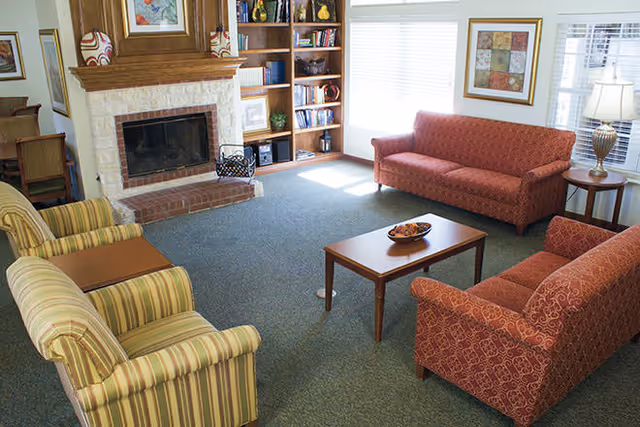 A cozy living room with two red patterned sofas, two striped armchairs, a wooden coffee table with a decorative bowl, a brick fireplace with a wooden mantle, built-in bookshelves filled with books and decor, and large windows letting in natural light.