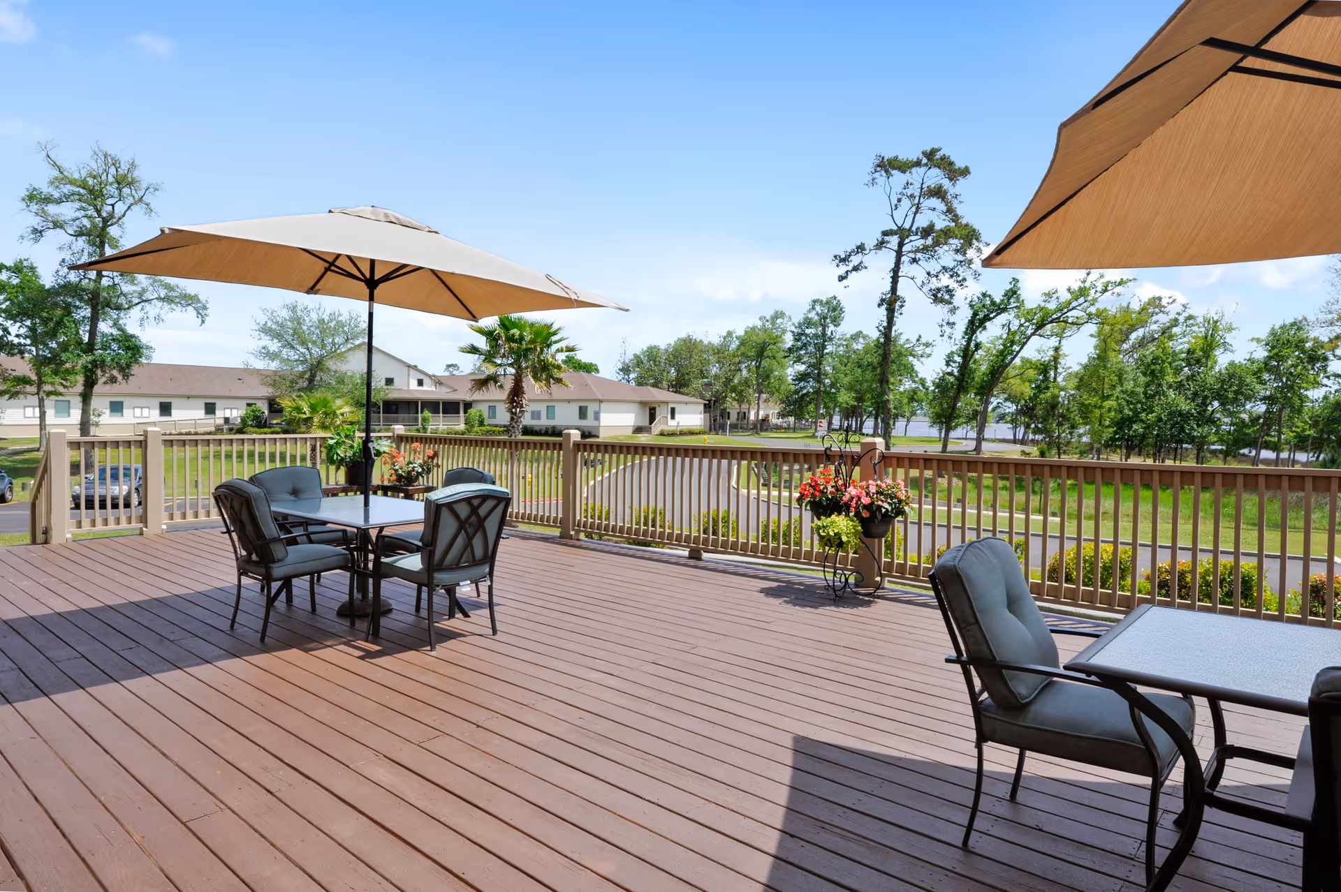 Outdoor wooden deck with two patio tables, each with cushioned chairs and large beige umbrellas providing shade. The deck overlooks a landscaped area with trees, a road, and buildings in the background under a clear blue sky.