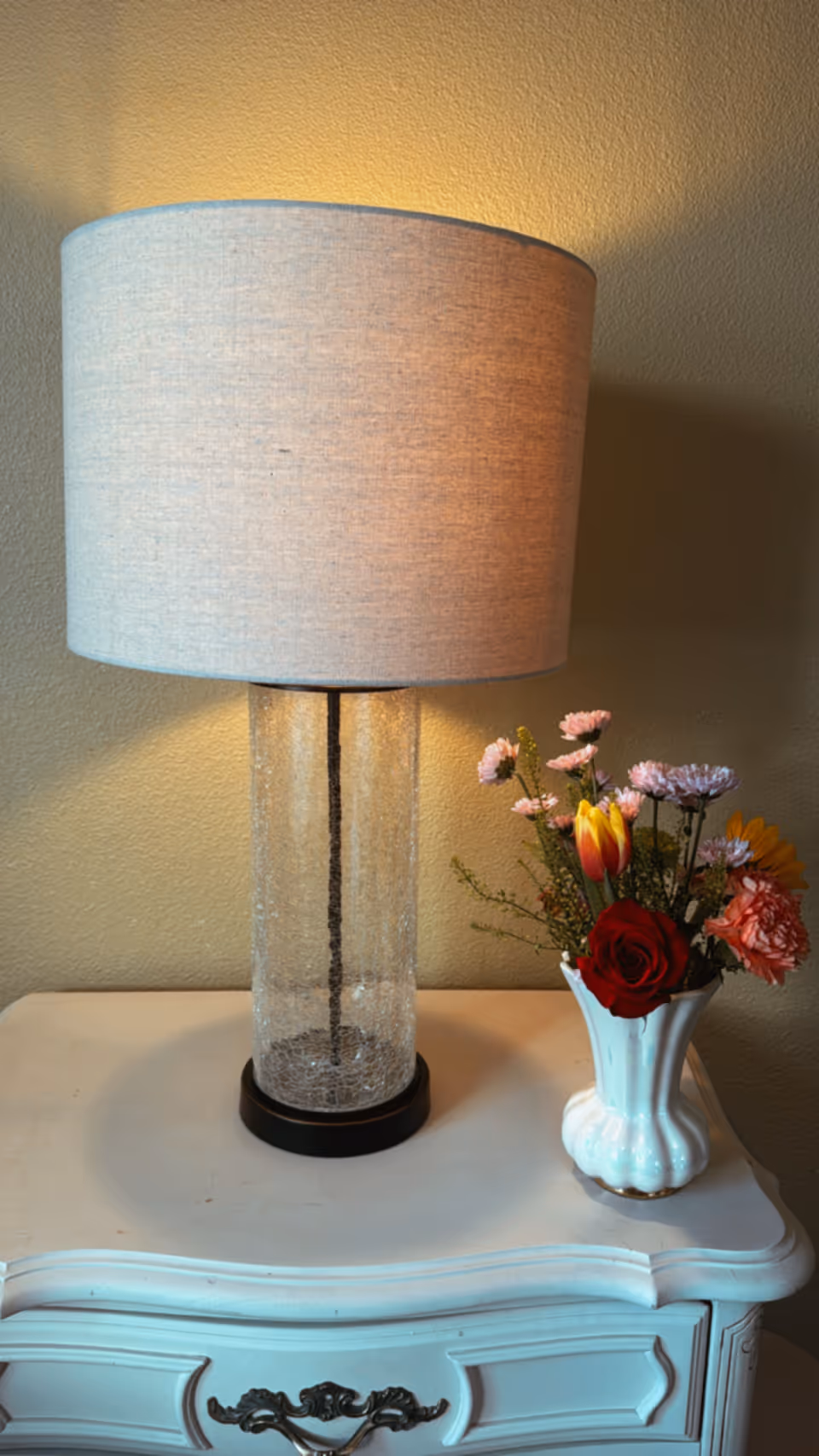 A lit table lamp with a textured glass base and a light gray fabric lampshade sits on a white vintage-style nightstand. Next to the lamp is a white ceramic vase holding a colorful bouquet of flowers including a red rose, yellow tulip, pink carnations, and other small blossoms. The background is a plain beige wall.