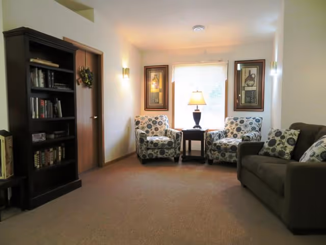 A cozy living room area with a brown sofa adorned with patterned cushions, two armchairs with floral upholstery, a small wooden side table with a lamp, a tall dark wooden bookshelf filled with books, and framed artwork on the walls. A window with a sheer curtain allows natural light into the room.