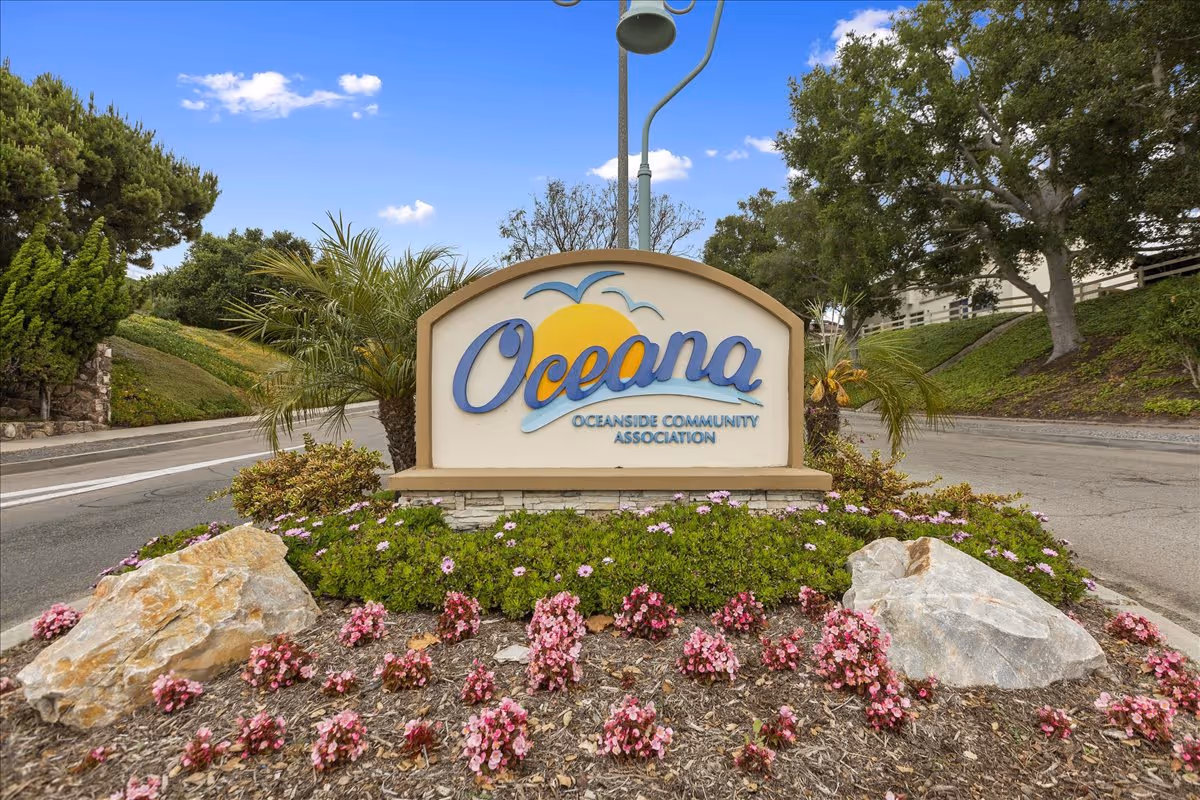 An entrance sign reading 'Oceana Oceanside Community Association' surrounded by landscaped flowers, rocks, and trees.