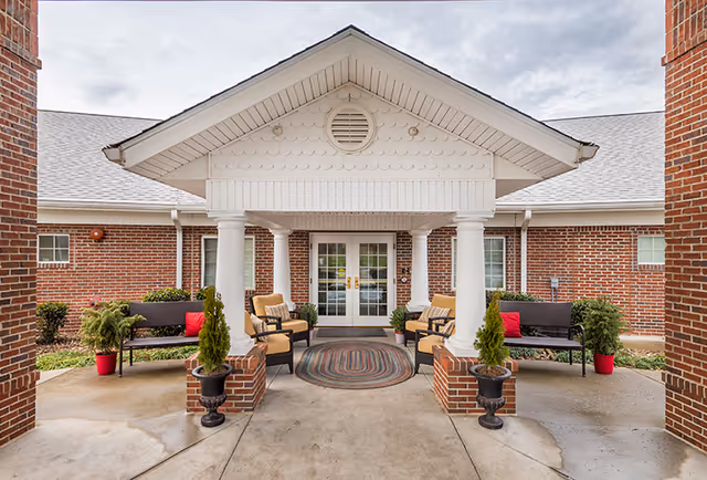 Entrance to a brick building with a covered porch supported by white columns. The porch area has outdoor seating including benches and cushioned chairs, with potted plants and a round rug in front of double glass doors.
