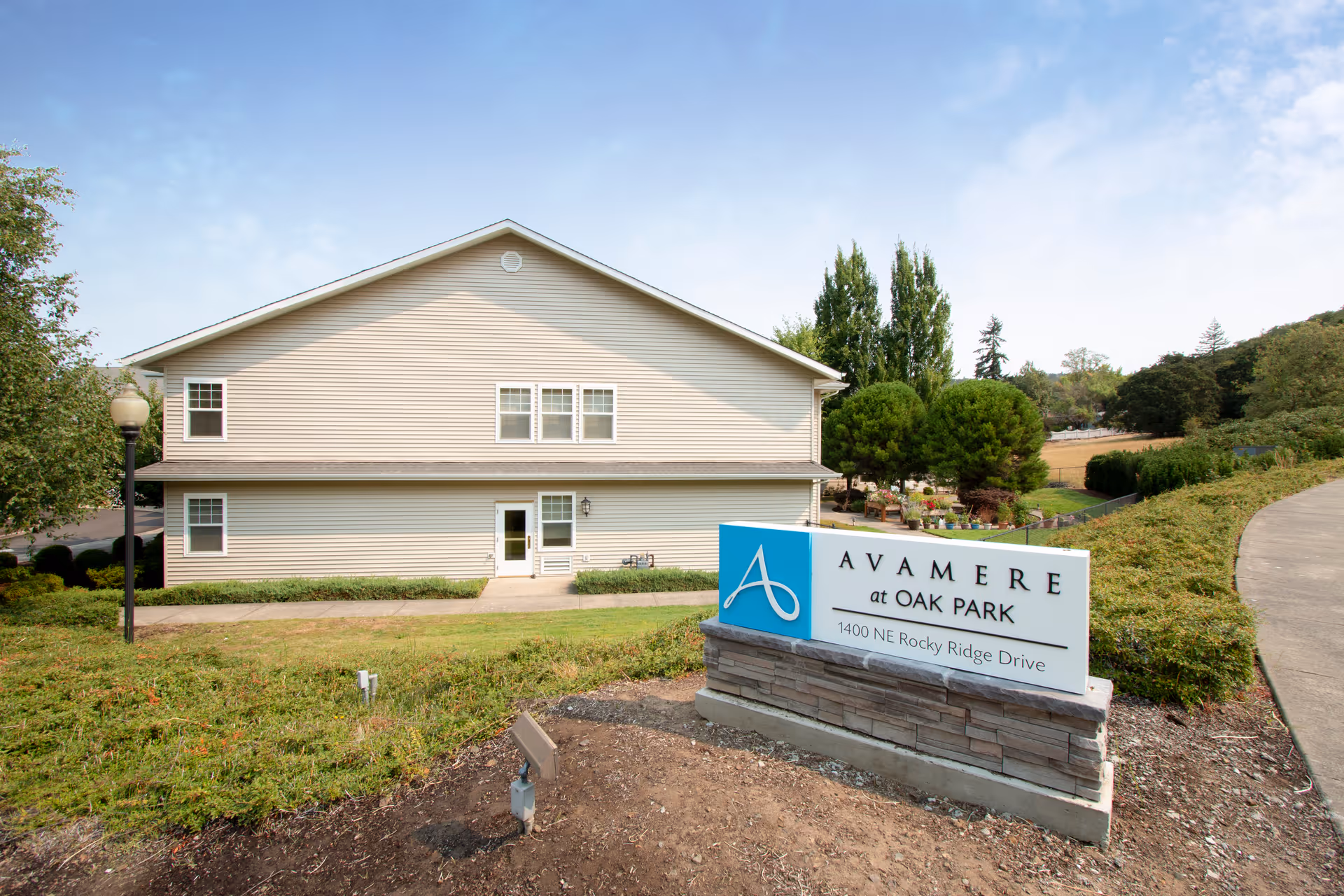 Front exterior of a two-story beige senior living building with landscaping and a sign reading 'Avamere at Oak Park, 1400 NE Rocky Ridge Drive'.