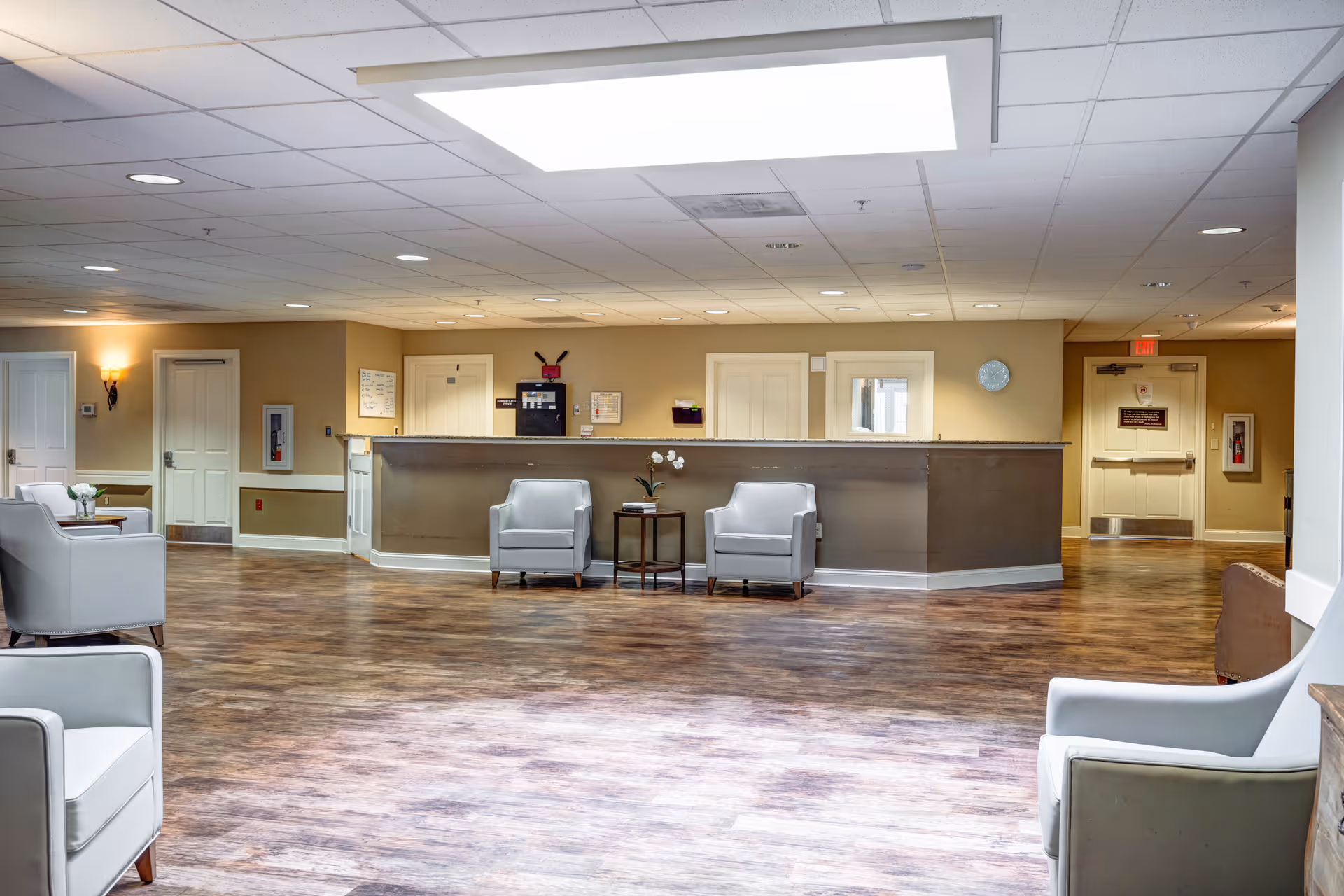 A spacious senior living facility common area with a reception desk in the background. The room features wood flooring, beige walls, and a white drop ceiling with recessed lighting. There are several light gray armchairs arranged around small tables with decorative plants. Doors and a fire extinguisher are visible along the walls.