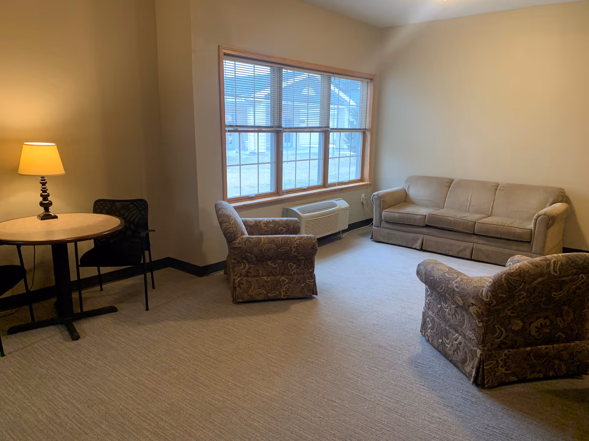 A cozy living room with a beige sofa, two patterned armchairs, a round table with two chairs, and a lit table lamp. A large window with blinds lets in natural light, and there is a wall-mounted air conditioning unit below the window.