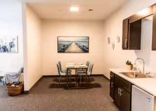 A small dining area with a table set for four, surrounded by four chairs. To the right is a kitchenette with a sink, countertop, and dark wooden cabinets. On the wall above the dining table is a framed picture of a wooden pier extending into the water. The room has neutral-colored walls and carpeted flooring.