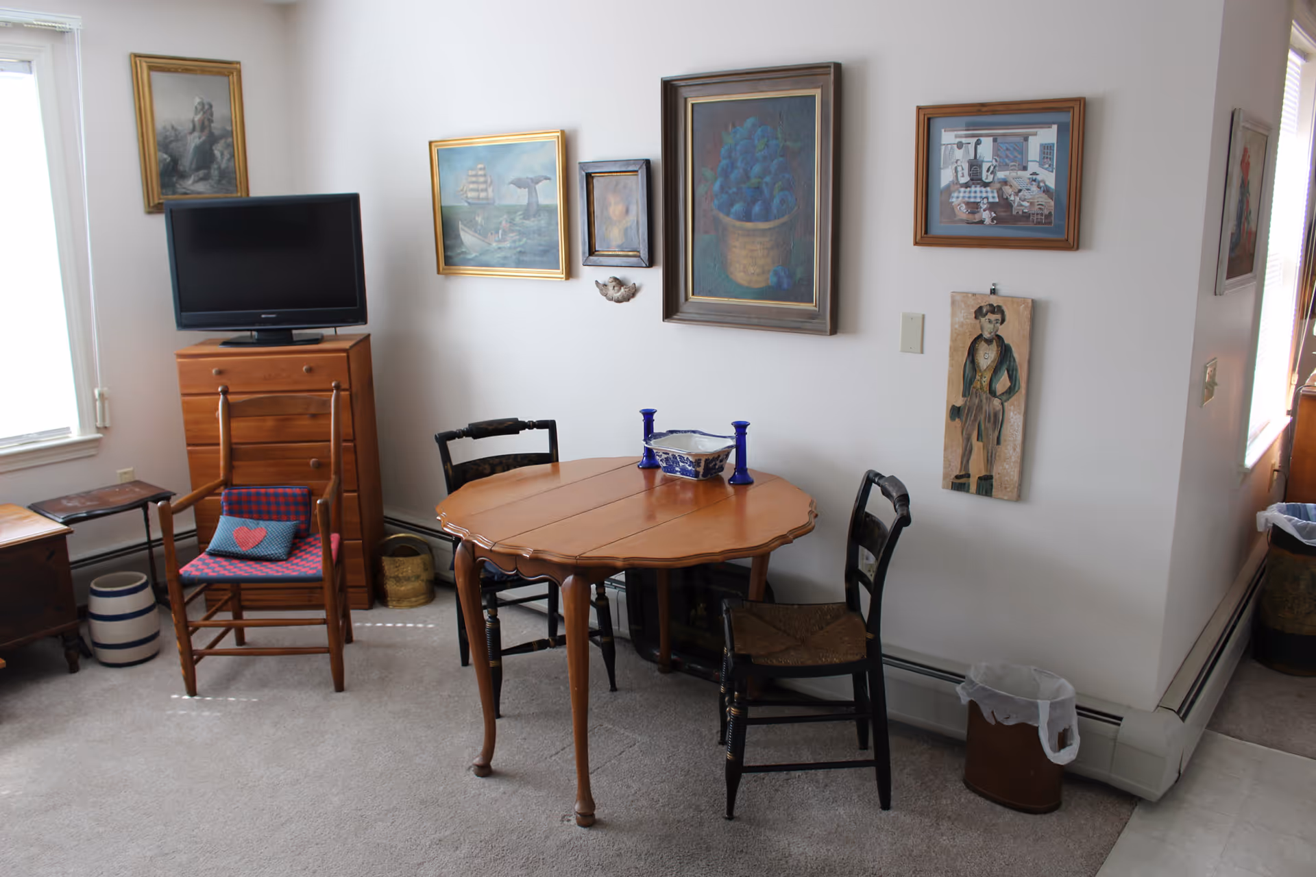 Small dining area with a round wooden table and chairs, a TV on a dresser, framed artwork on white walls, and a window.