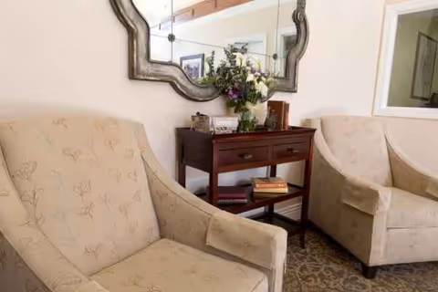 Cozy senior living lounge with two beige armchairs, a console table, flowers, and a wall mirror.