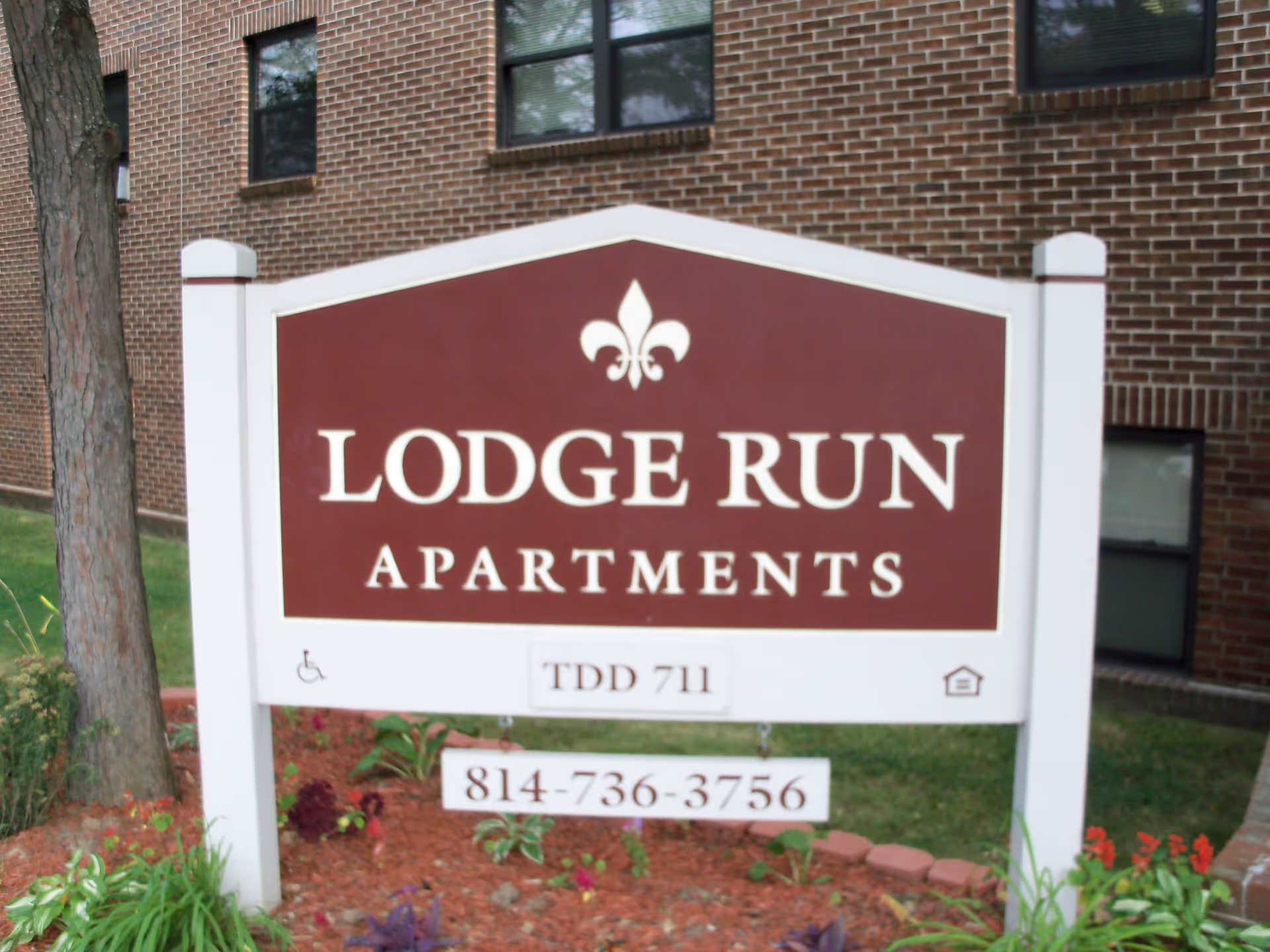 A maroon and white sign for Lodge Run Apartments in front of a brick building with windows. The sign includes a phone number and accessibility symbols, surrounded by landscaping with plants and mulch.