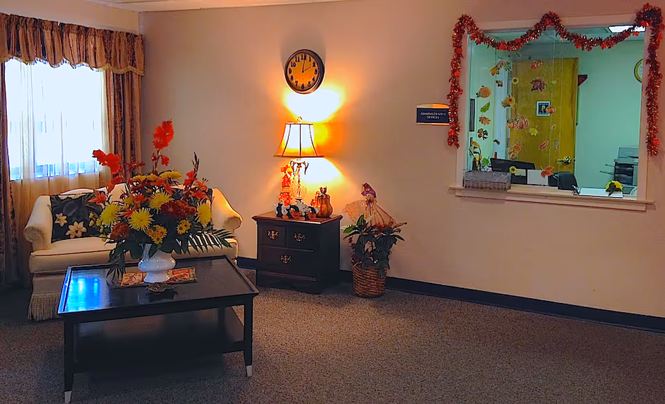 A cozy interior room with a white couch adorned with floral pillows, a dark wooden coffee table holding a large vase of colorful flowers, and a side table with a lamp and decorative items. A clock is mounted on the wall above the lamp. There is a window with autumn-themed decorations looking into an office space with a yellow door and office furniture.