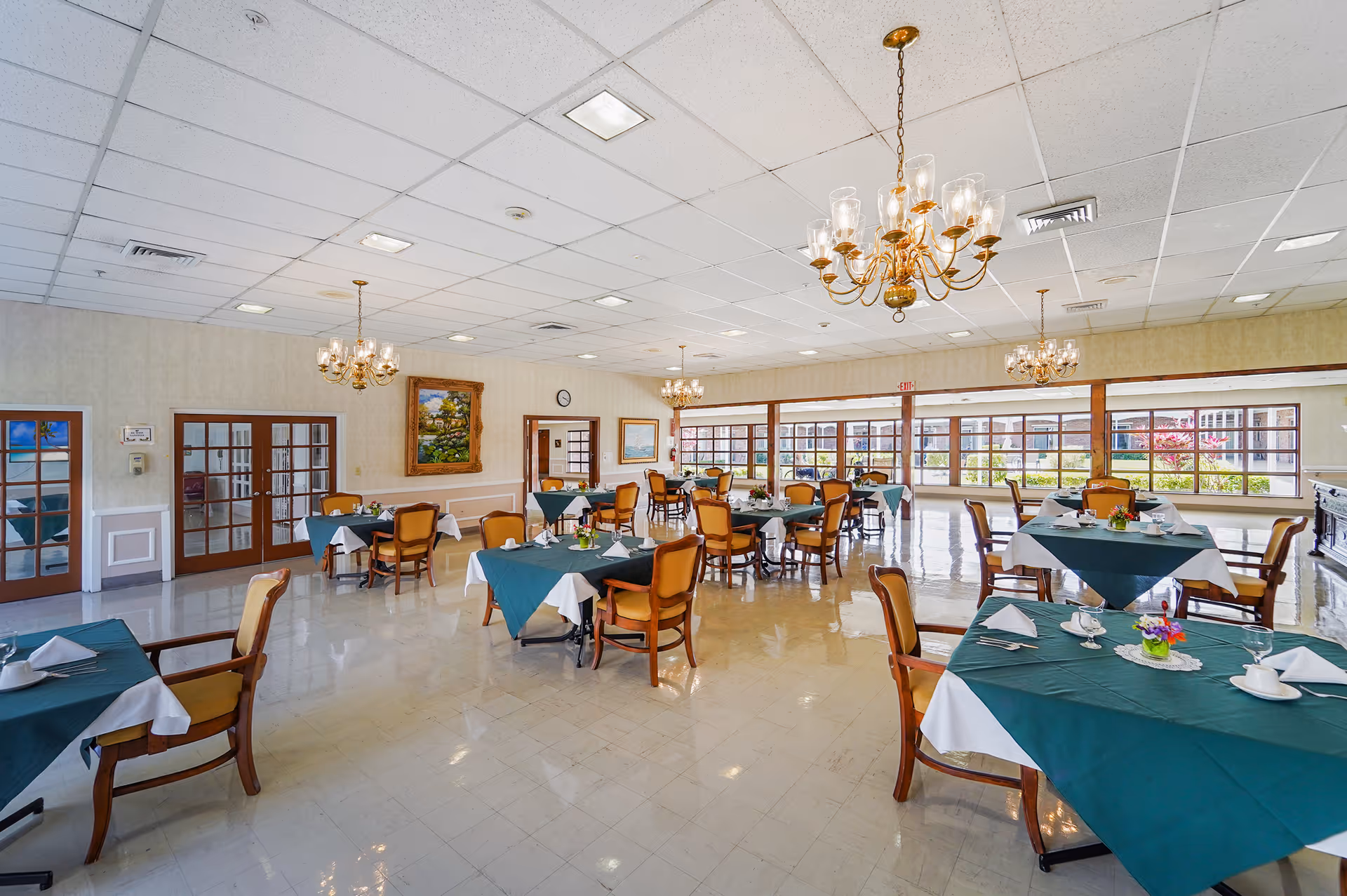 A spacious dining room with multiple tables covered in green tablecloths and white napkins, surrounded by wooden chairs with yellow cushions. The room features large windows letting in natural light, chandeliers hanging from the ceiling, and paintings on the walls.