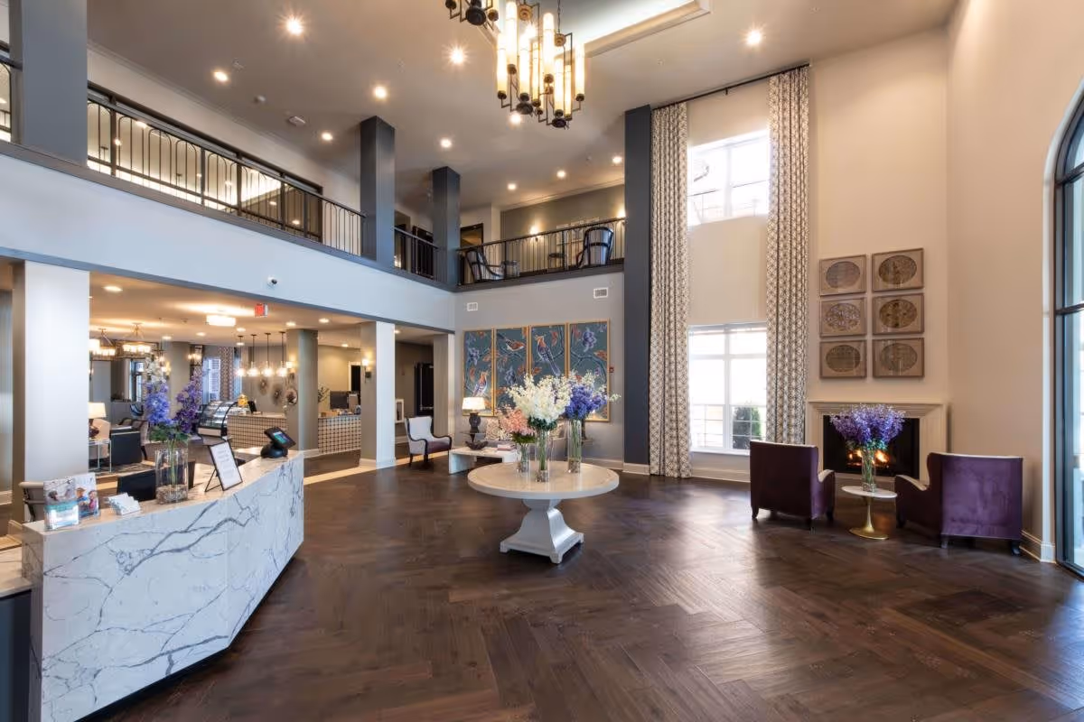 Spacious and elegant senior living facility lobby with high ceilings, a chandelier, large windows with patterned curtains, a round table with flower arrangements in the center, a marble reception desk on the left, and a seating area with two purple chairs and a small table with flowers near a fireplace on the right.