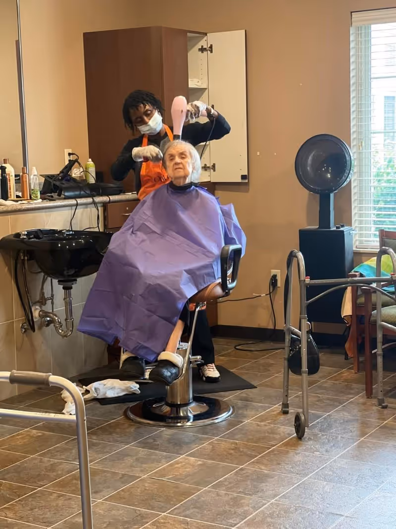 An elderly woman wearing a purple cape is seated in a salon chair while a hairstylist wearing an orange apron and face mask blow-dries her hair. The room has tiled floors, a hair washing station, a walker, and a window with blinds letting in natural light.