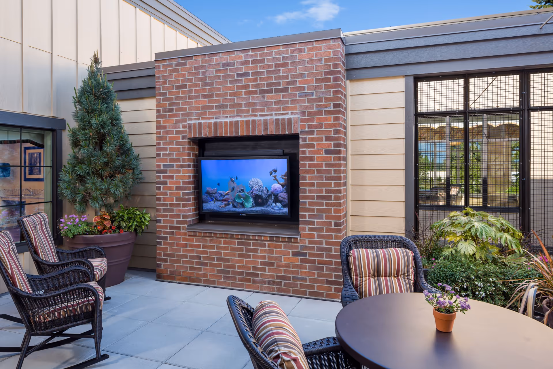 Outdoor patio area with a brick fireplace that has a flat-screen TV mounted inside it. The patio has cushioned wicker chairs with striped cushions around a round table with a small potted plant. There are large potted plants and greenery around the patio, and a window with a black grid frame is visible on the right side.