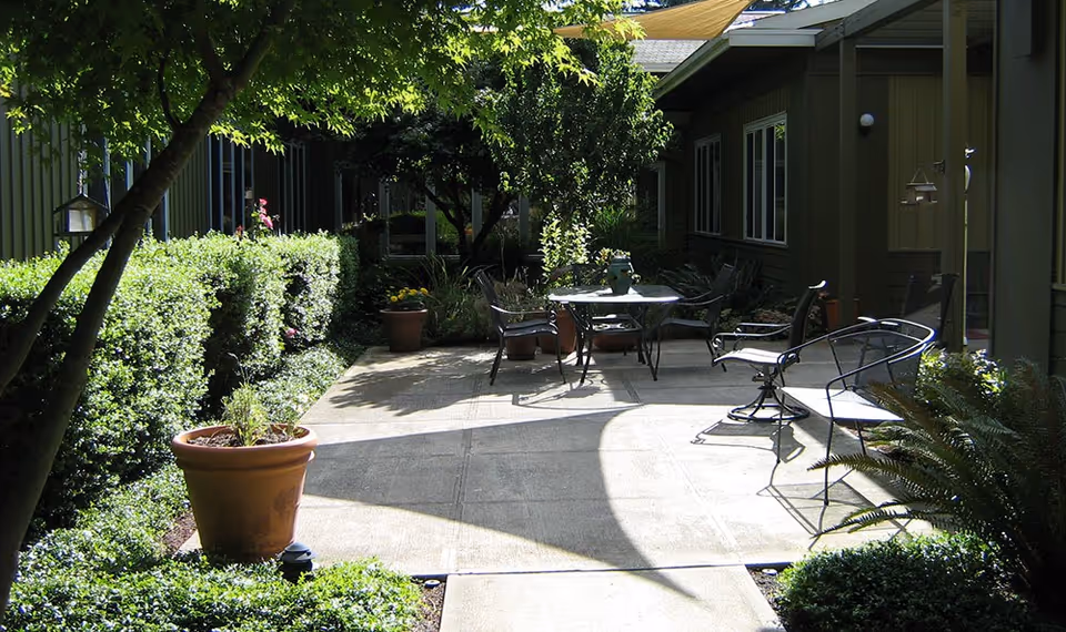 Outdoor patio area at Maple Grove Memory Care with metal chairs and tables, surrounded by green bushes, potted plants, and trees providing shade.