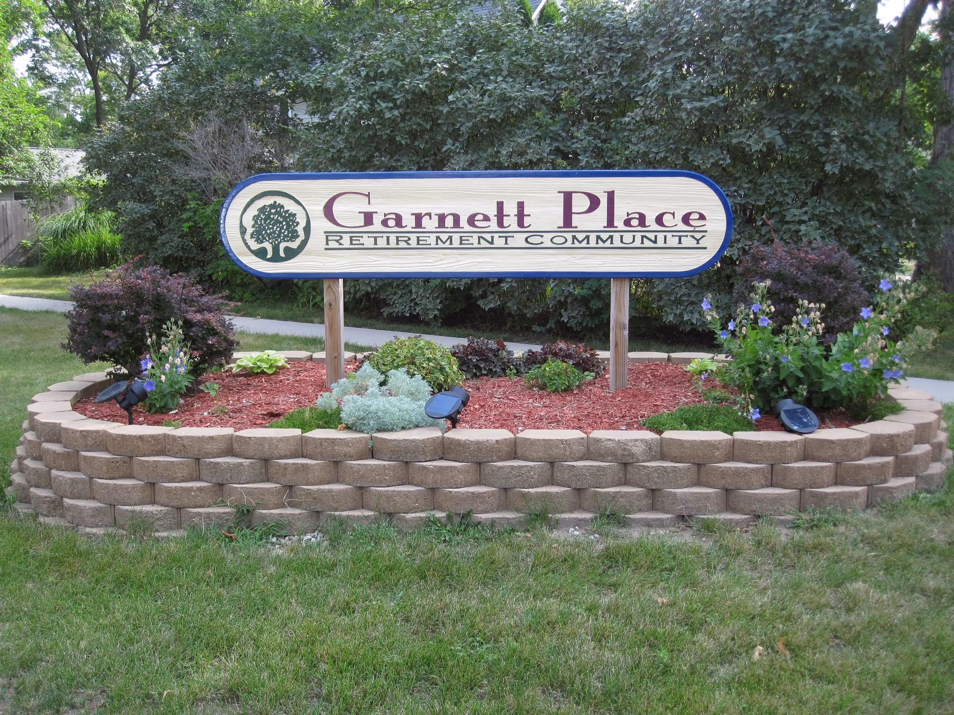A wooden sign reading "Garnett Place Retirement Community" mounted in a landscaped flowerbed with shrubs and flowers.