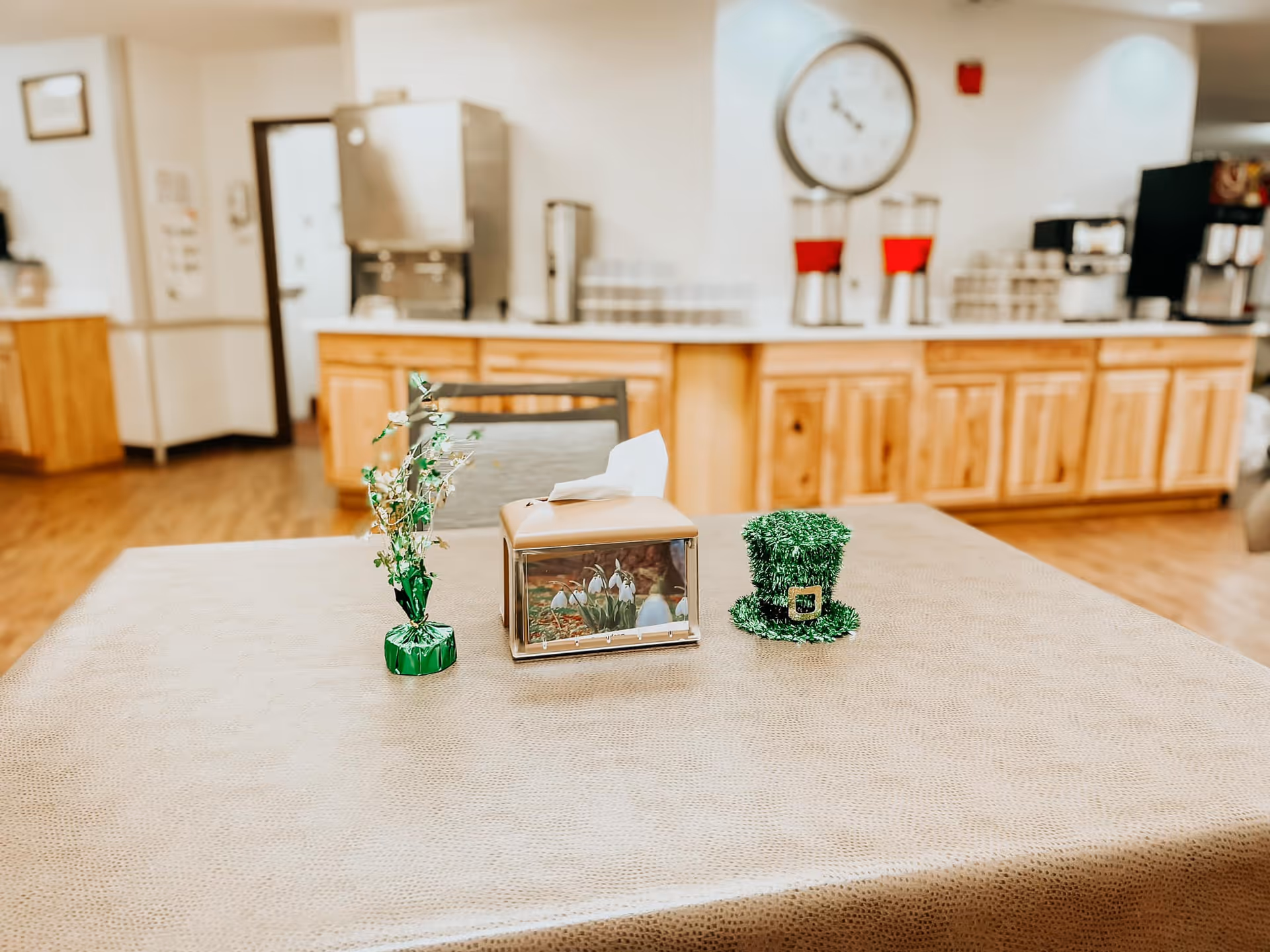 Table with a tissue box and small green decorations in a dining/common area with a serving counter and beverage dispensers in the background.