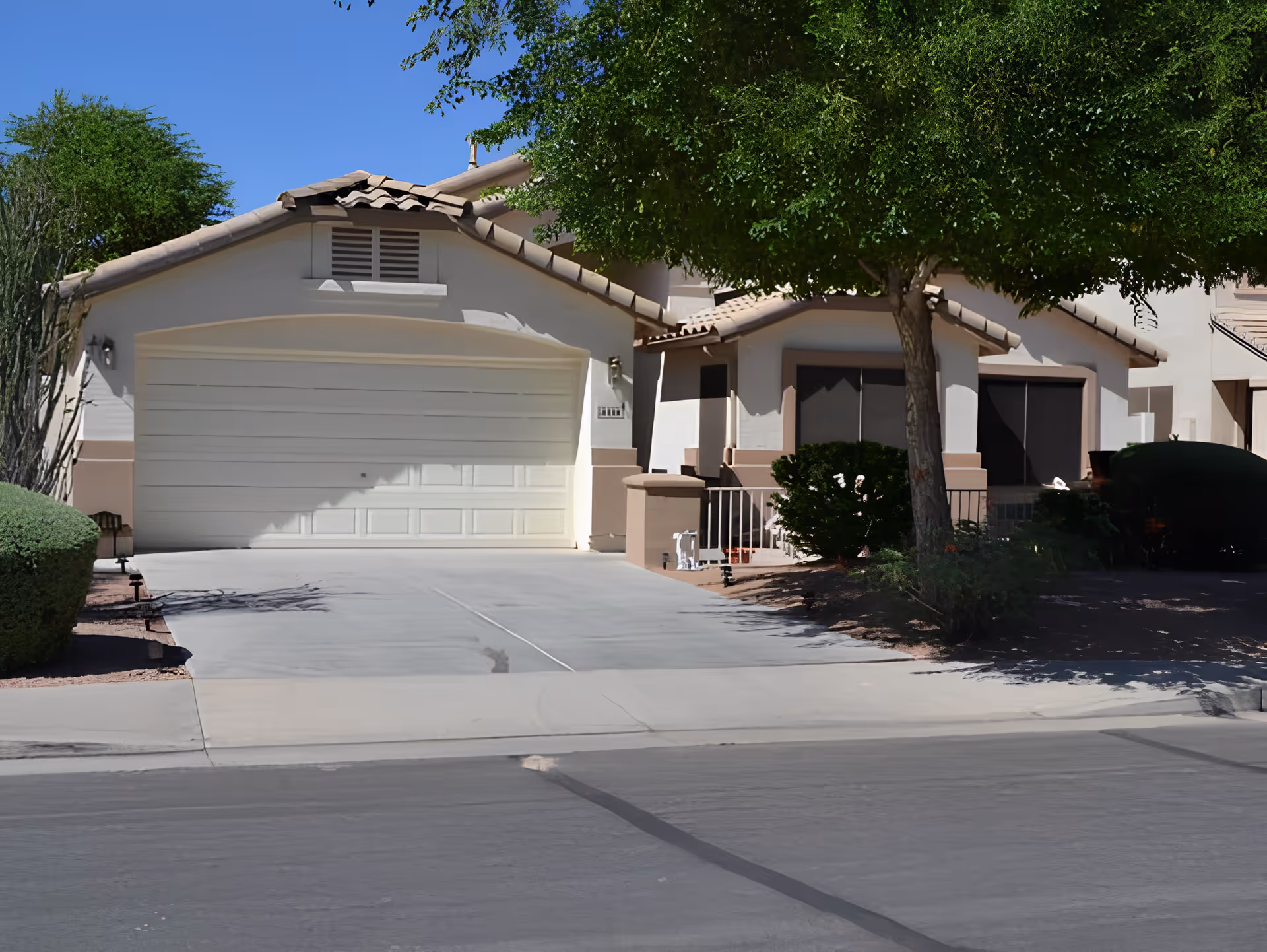 Front exterior view of a single-story house with a closed white garage door, a driveway, and a tree partially shading the front yard. The house has a tiled roof and beige walls with brown trim.