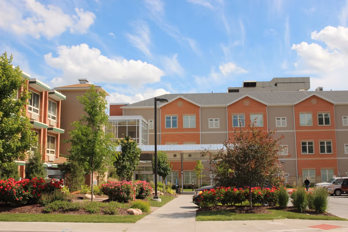 Exterior view of Ambassador Health of Omaha building with a landscaped garden featuring trees, bushes, and flowers in front. The building has multiple windows and a covered entrance. Cars are parked on the right side under a partly cloudy blue sky.