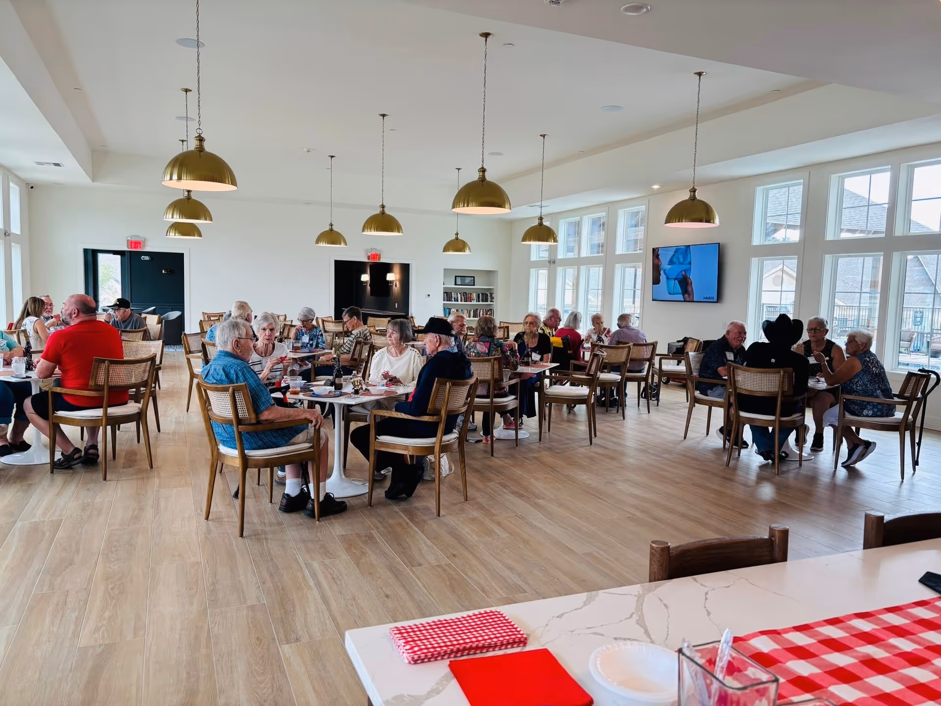 A bright and spacious dining room with large windows letting in natural light. Several elderly people are seated at round tables, engaging in conversation and enjoying their meals. The room features wooden flooring, modern pendant lights hanging from the ceiling, and a television mounted on the wall. The atmosphere appears lively and social.