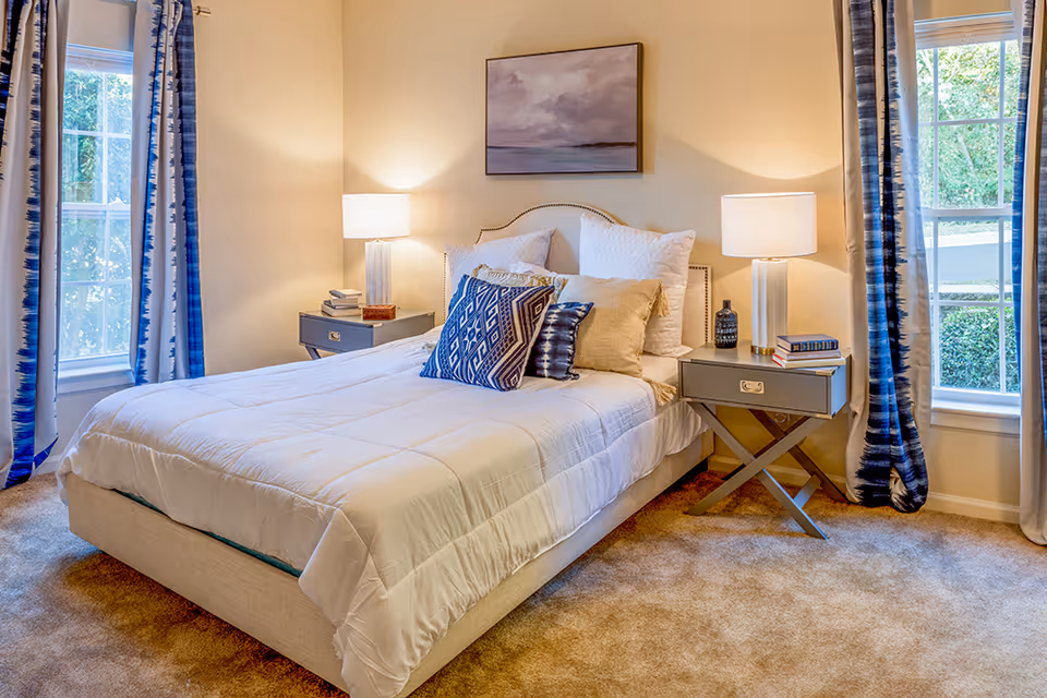A cozy bedroom with a neatly made bed featuring white bedding and multiple decorative pillows in blue and beige tones. There are two bedside tables, each with a lamp, books, and decorative items. The room has beige walls, carpeted floor, two windows with blue and white patterned curtains, and a framed artwork hanging above the bed.