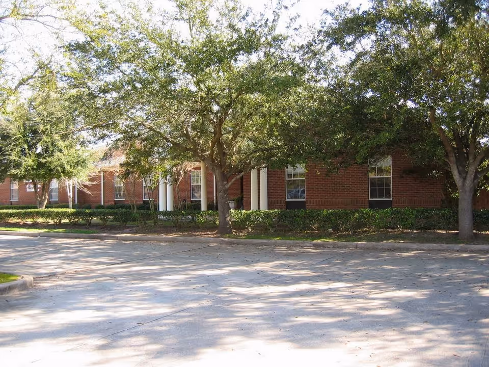 Brick one-story building with white columns partially obscured by trees and shrubs and a paved parking area in front.