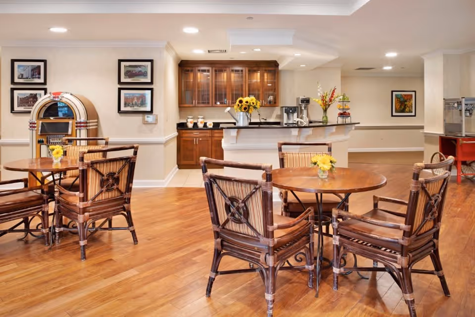 A cozy common area with wooden floors featuring two round wooden tables each surrounded by four wicker chairs with cushions. Each table has a small vase with yellow flowers. In the background, there is a counter with a coffee machine, a vase with flowers, and cabinets with glass doors. On the left side, there is a vintage jukebox and framed pictures on the wall. On the right side, there is a popcorn machine on a red stand and more framed artwork on the wall.