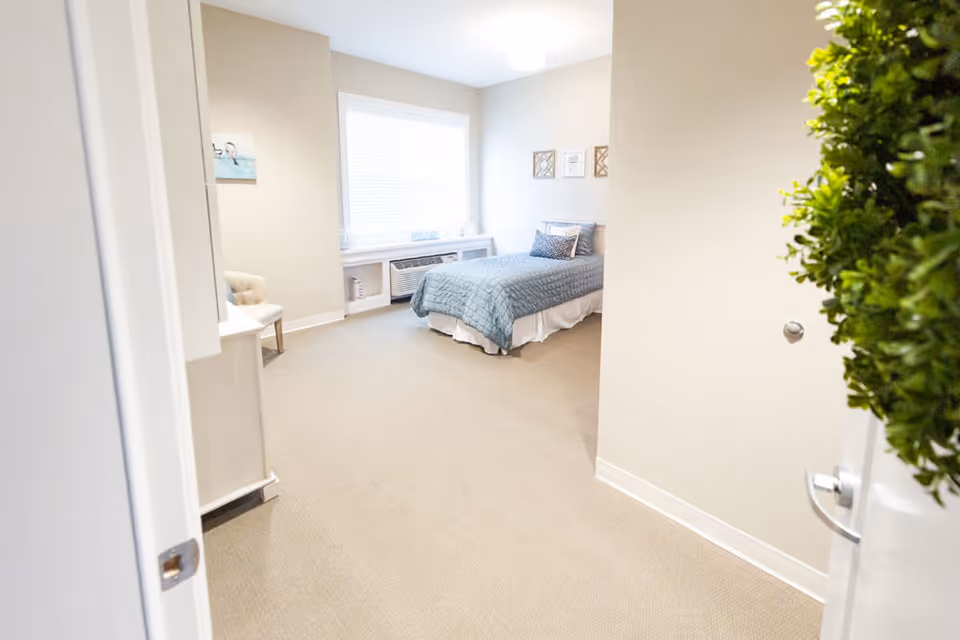 View into a senior living bedroom with a single bed covered in a blue quilt, a window with blinds, a small chair, and light beige walls and carpet. A green wreath is partially visible on the door.