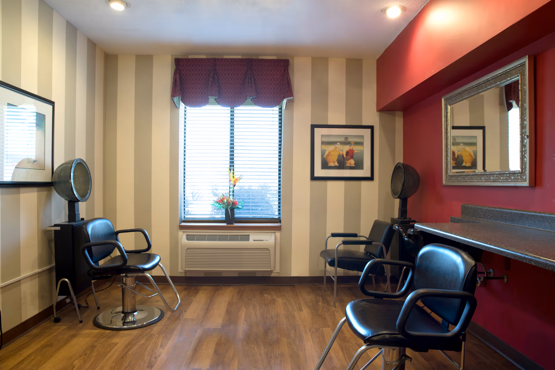 Interior of a salon room with three black salon chairs, two hair dryers, a large mirror on a red wall, striped beige wallpaper, a window with blinds and a red valance, and a small flower arrangement on the windowsill.