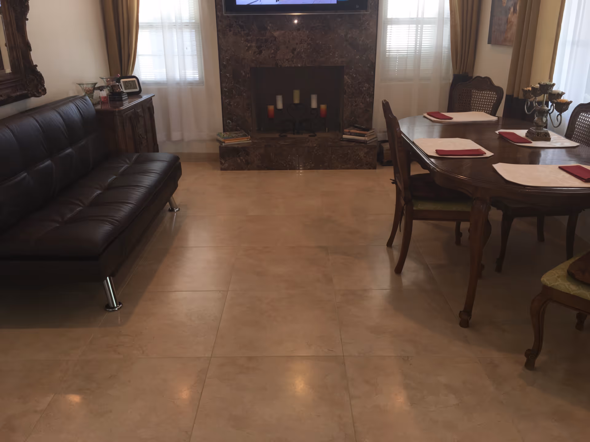 Interior room with a dark brown leather sofa on the left, a wooden dining table with four chairs on the right, each place set with a placemat and red napkin. In the center background is a marble fireplace with candles and books on the hearth, flanked by two windows with sheer white curtains and beige drapes.