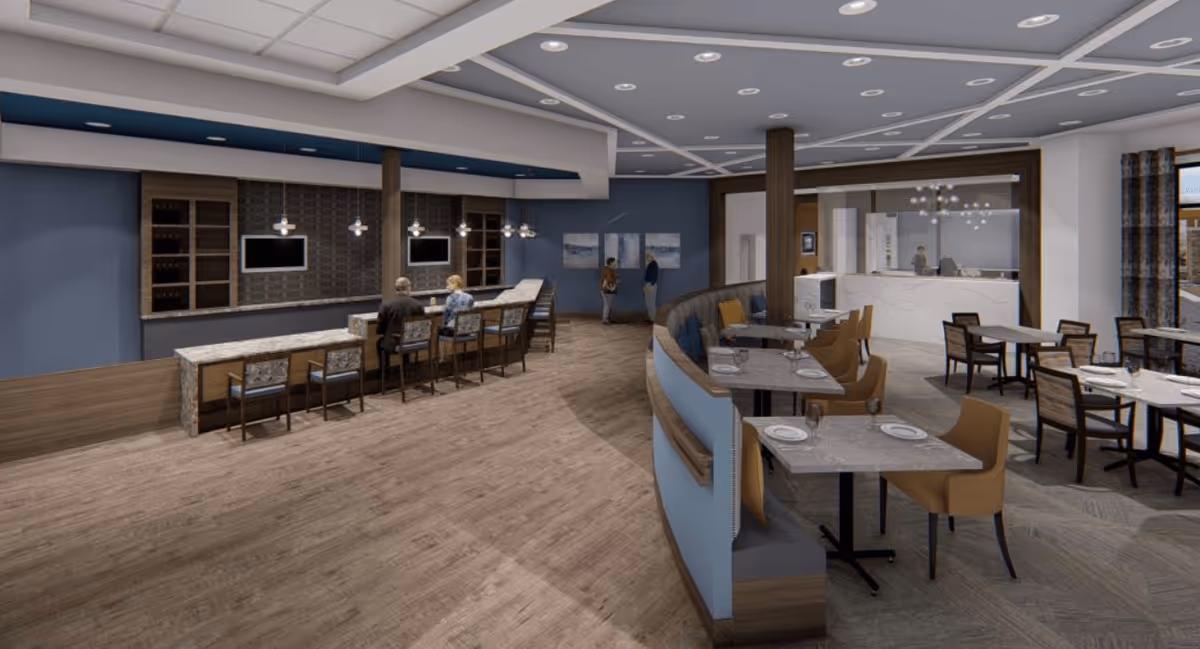 Interior view of a modern dining area in a senior living facility with a bar counter on the left where two people are seated, and several dining tables with chairs and place settings on the right. The space features wood flooring, blue and neutral tones, pendant lighting, and a reception desk in the background.