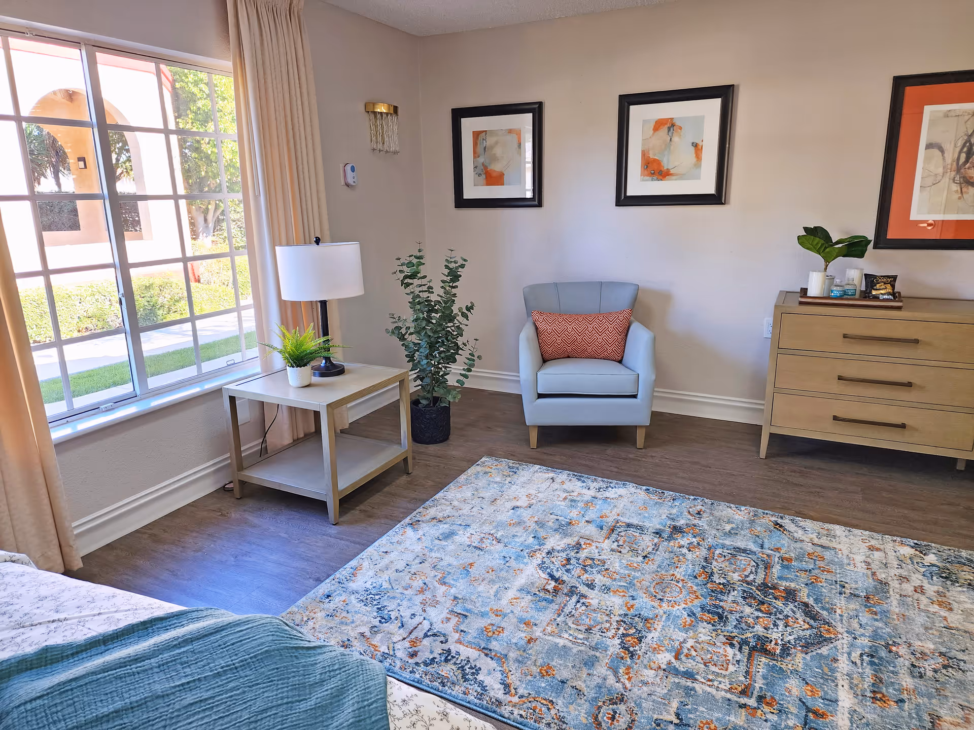 A cozy room with a large window letting in natural light, beige curtains, a light blue armchair with an orange patterned pillow, a wooden side table with a lamp and a small plant, a large decorative rug with blue and orange patterns, a wooden dresser with a plant and framed artwork on the wall above.