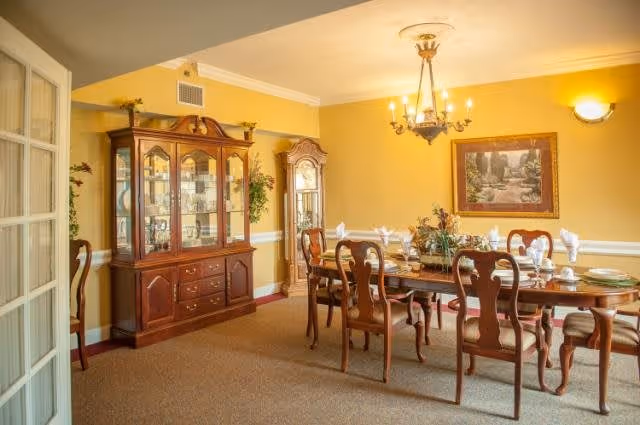 A formal dining room with a wooden dining table set for eight, featuring folded white napkins and glassware. The room has yellow walls, a chandelier hanging from the ceiling, a large wooden china cabinet filled with glassware, a grandfather clock, and a framed painting on the wall.