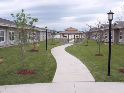 Curved concrete walkway through a grassy courtyard between single-story buildings leading to a central gazebo with lamp posts and young trees.