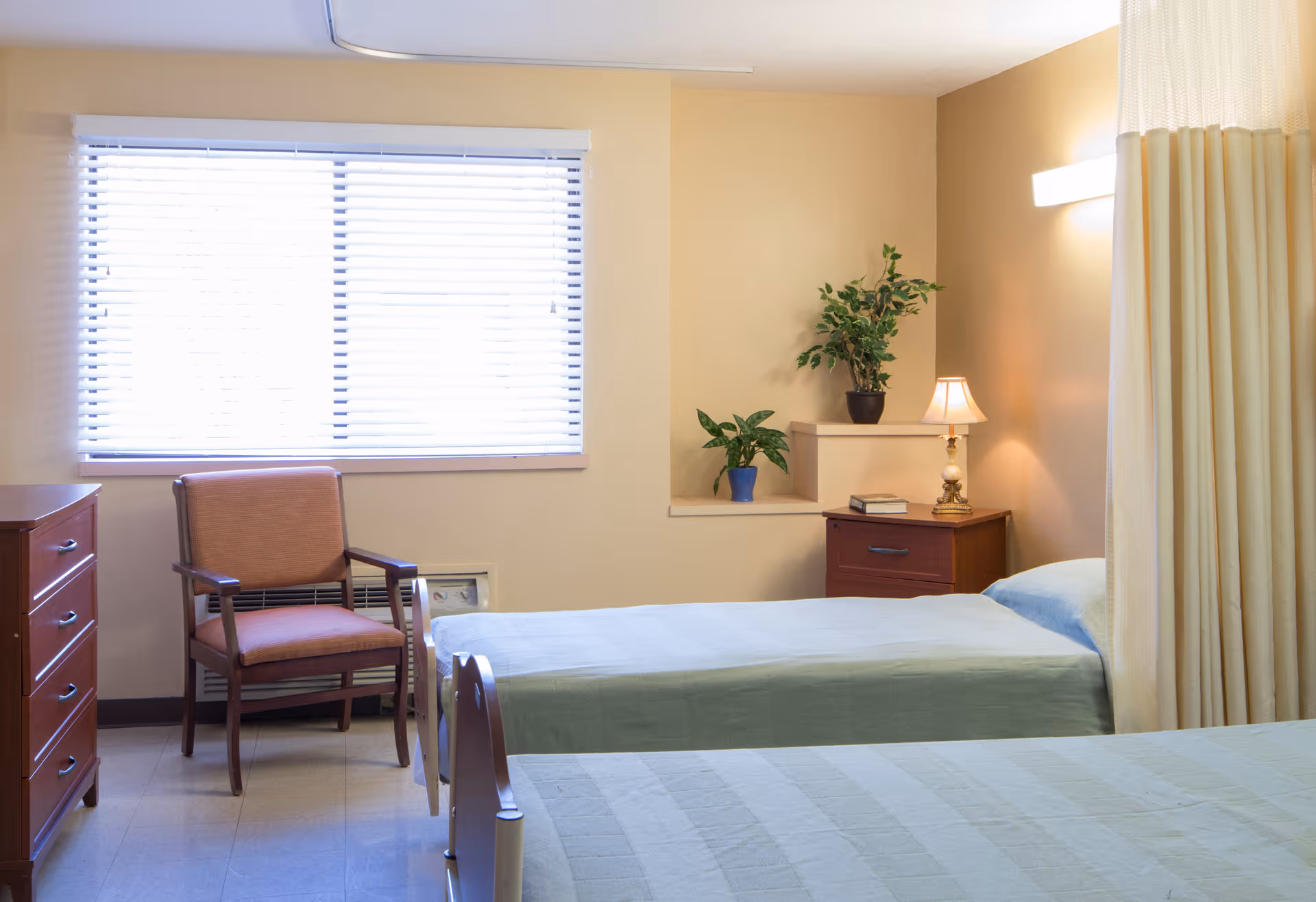 A simple bedroom in a senior living facility with two single beds covered in light green bedspreads. There is a wooden chair with armrests near a window with white blinds. A wooden dresser and a nightstand with a small lamp and two potted plants are also visible. A beige privacy curtain is partially drawn on the right side of the image.